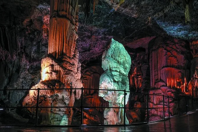 Angestrahlte Stalagmiten und eine helle Säule neben einem Fußweg mit Geländer in einer dunklen Höhle