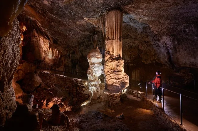 Ein hoher orangefarbener Stalagmit, beleuchtet in einer dunklen Höhlenkammer, ein Besucher von hinten auf dem Weg